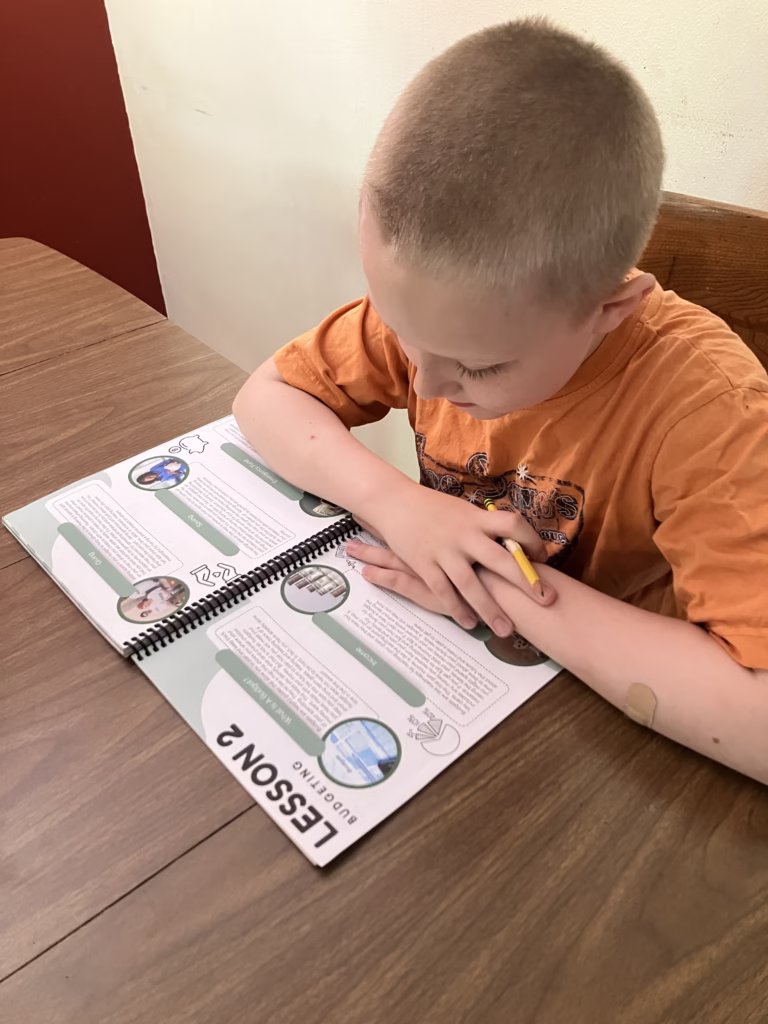 Personal finance is an important life skill to teach your homeschoolers. Beyond Personal Finance offers kids real practice in this skill. A child reads Lesson 2: Budgeting inside of the Before Personal Finance student workbook.
