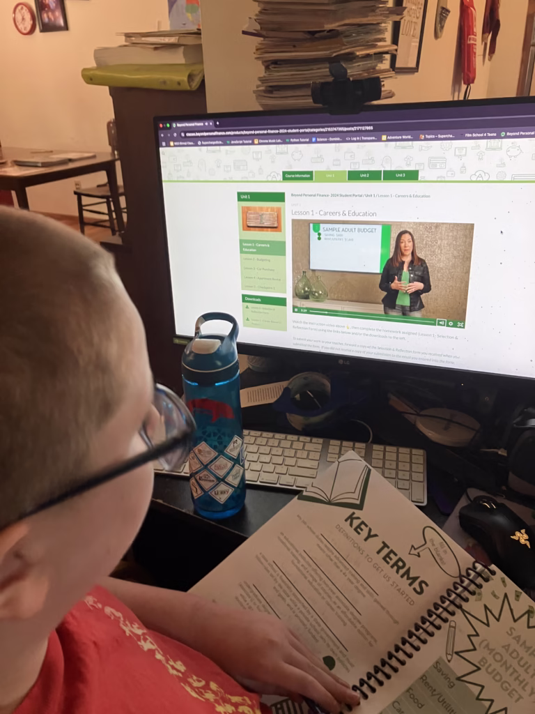 Personal finance is an important life skill to teach your homeschoolers. Beyond Personal Finance offers kids real practice in this skill. A child watches a lesson from Beyond Personal Finance on the computer and works on Key Terms inside the included student workbook. 